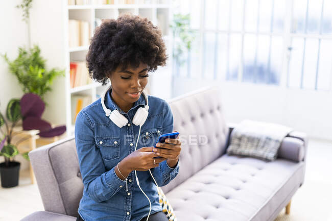 Жінка з навушниками, використовуючи мобільний телефон, сидячи на дивані вдома — стокове фото