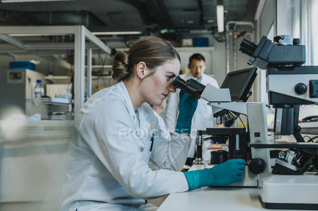 Молодая женщина анализирует микроскоп человеческого мозга под микроскопом, сидя с учеными на заднем плане в лаборатории — стоковое фото