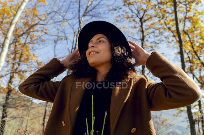 Donna sorridente indossando cappello guardando lontano mentre in piedi nella foresta — Foto stock