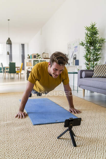 Молодой человек смотрит мобильный телефон во время занятий на коврике дома — стоковое фото