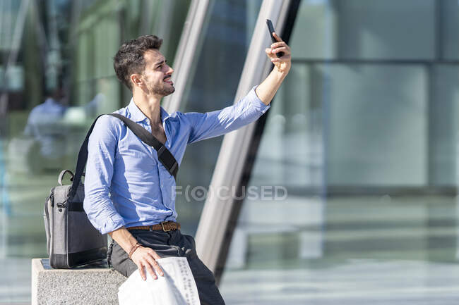 Hombre de negocios sonriente tomando selfie a través del teléfono móvil mientras está sentado al aire libre - foto de stock