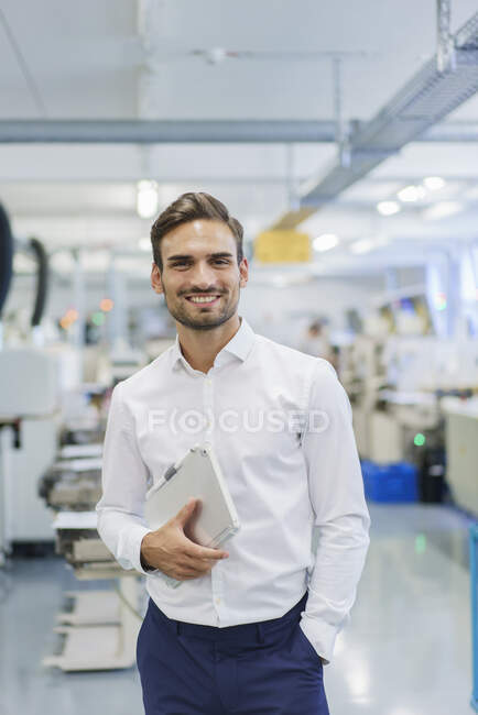 Усміхнений молодий бізнесмен тримає цифровий планшет, стоячи руками в кишенях на освітленій фабриці — стокове фото