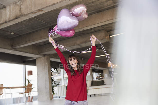 Mujer madura alegre sosteniendo el globo de helio en forma de corazón mientras está sentado en casa - foto de stock