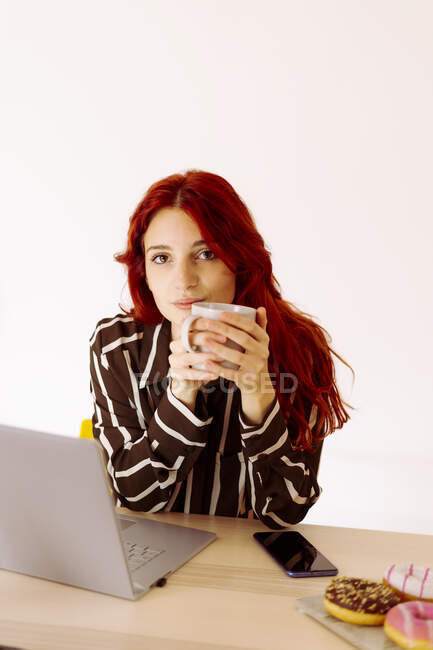 Rothaarige Geschäftsfrau trinkt Kaffee, während sie am Tisch im Büro sitzt — Stockfoto
