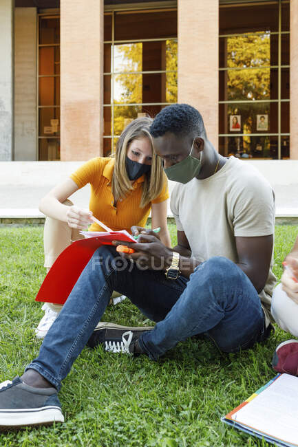 Männliche und weibliche Freunde sitzen auf Gras, während sie gemeinsam auf dem Universitätscampus studieren — Stockfoto