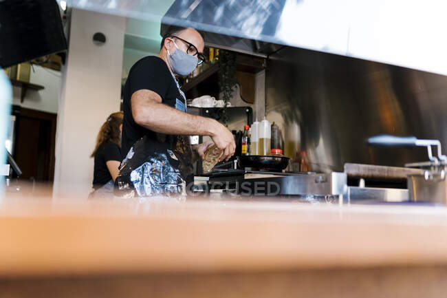Мужчина бариста носить защитную маску для лица во время приготовления пищи на кухне кофе-бара во время COVID-19 — стоковое фото