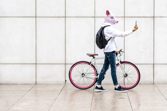 Hombre joven con bicicleta con máscara de cerdo mientras toma selfie en el teléfono móvil - foto de stock