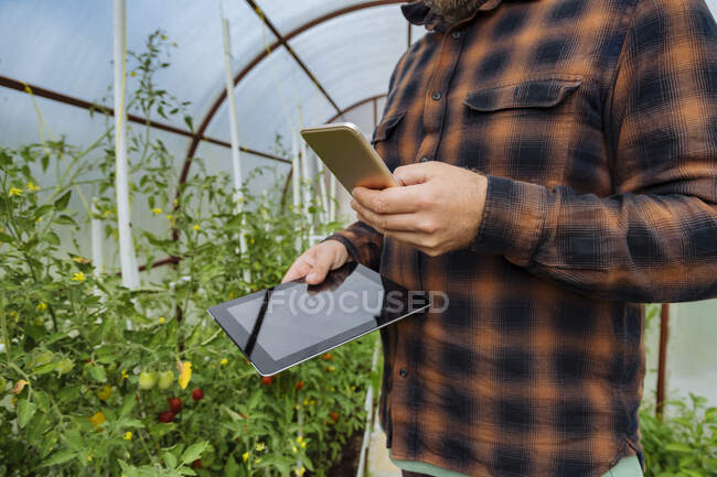 Homme tenant tablette numérique tout en utilisant le téléphone mobile à la serre — Photo de stock