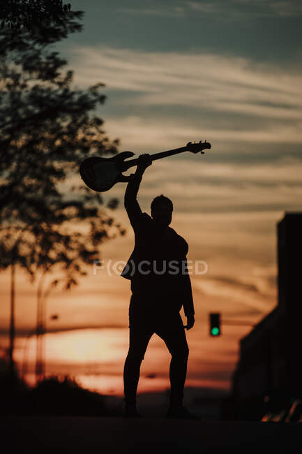 Музыкант, держащий бас-гитару, стоя на дороге во время заката — стоковое фото