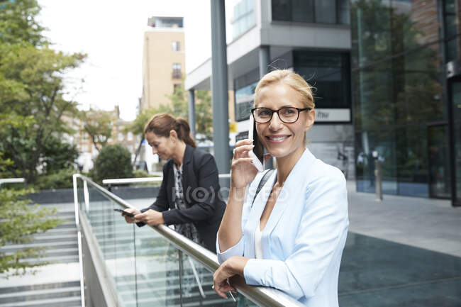 Улыбающаяся женщина разговаривает по мобильному телефону, в то время как коллега стоит на заднем плане в городе — стоковое фото