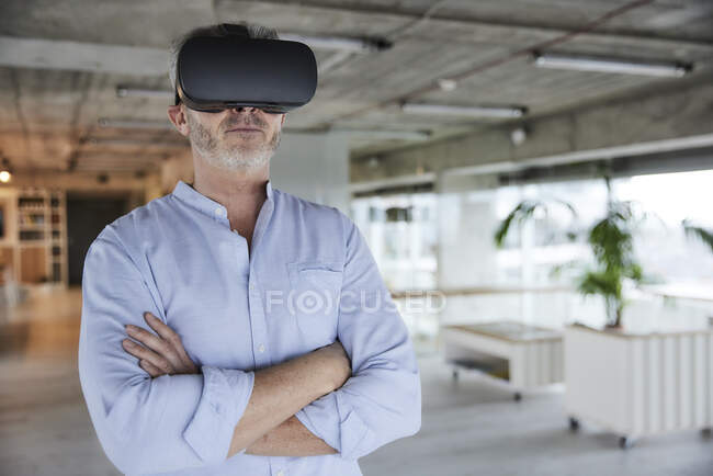 Зрелый бизнесмен, использующий гарнитуру виртуальной реальности, стоя со скрещенными руками дома — стоковое фото
