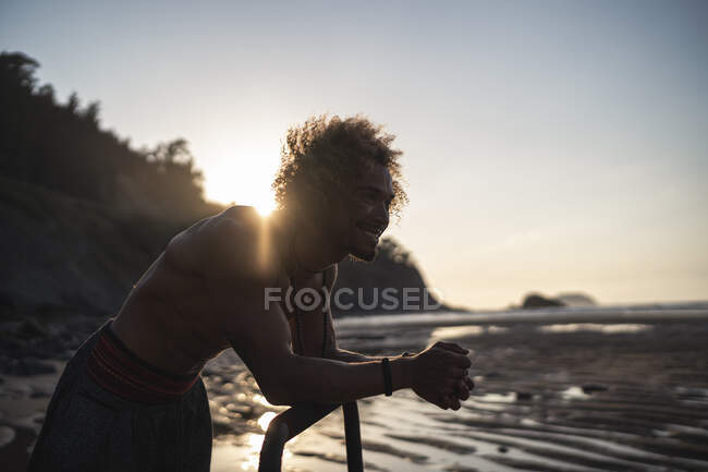 Shirtless young man looking away while leaning on parallel bars at beach — Stock Photo