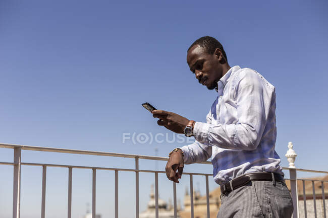 Imprenditore maschile utilizzando smartphone mentre appoggiato su ringhiera contro cielo blu chiaro — Foto stock