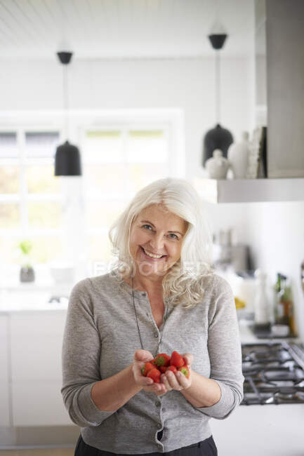 Усміхаючись, жінка на пенсії тримає свіжу полуницю, стоячи вдома на кухні. — стокове фото