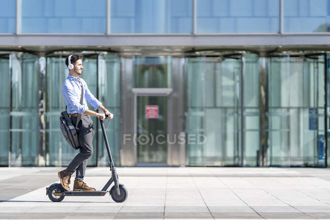 Junger Geschäftsmann mit Kopfhörer fährt Roller auf Fußweg — Stockfoto