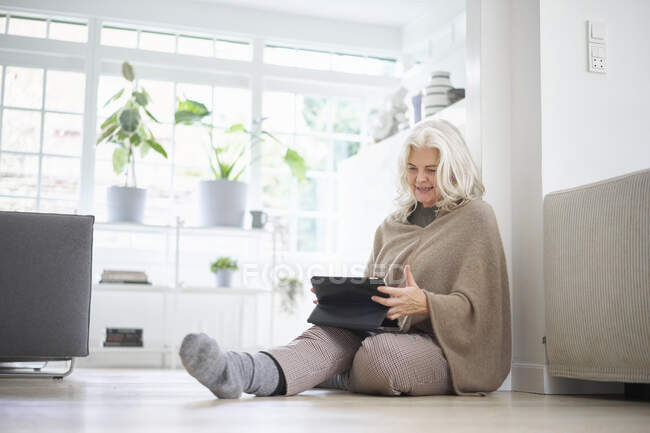 Mujer jubilada mayor sentada en el suelo usando tableta digital en la sala de estar en el apartamento - foto de stock