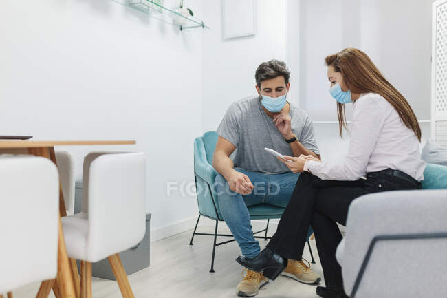 Médico y paciente usando mascarilla facial usando tableta digital mientras está sentado en el consultorio - foto de stock