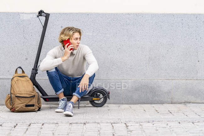 Homem com mochila falando no telefone celular enquanto sentado em scooter elétrico empurrar contra a parede — Fotografia de Stock