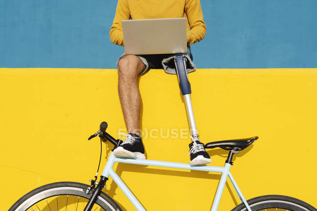 Homme avec un membre artificiel et le pied en utilisant un ordinateur portable tout en étant assis sur un mur multicolore — Photo de stock