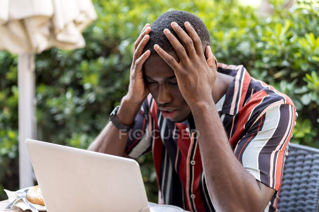 Напряженный молодой человек сидит с головой в руках, глядя на ноутбук в кафе — стоковое фото