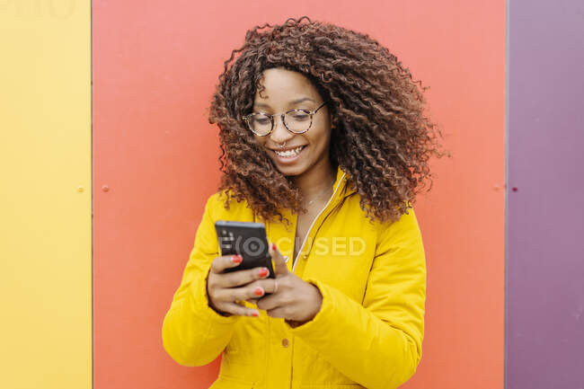 Mujer joven usando chaqueta usando teléfono móvil mientras está de pie contra la pared multicolor - foto de stock