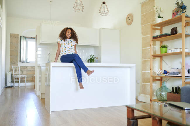 Sonriente joven afro mujer sentada en el mostrador de la cocina en casa - foto de stock