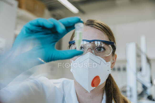 Молодая женщина в защитной маске и очках осматривает пробирку в лаборатории — стоковое фото