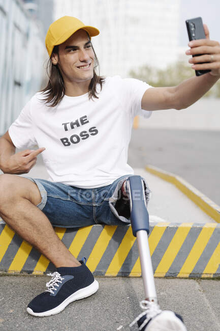 Hombre con extremidad artificial y pie tomando selfie mientras está sentado en la carretera en la ciudad - foto de stock