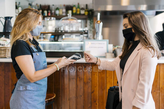 Безробітна жінка в масці перед обличчям платить кредитною карткою в кафе під час коронавірусної кризи. — стокове фото