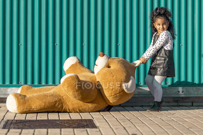 Mädchen schleift Teddybär auf Fußweg gegen grüne Wand — Stockfoto