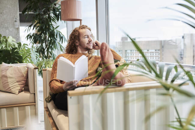 Вдумчивый человек смотрит в окно держа книгу, сидя дома на диване — стоковое фото