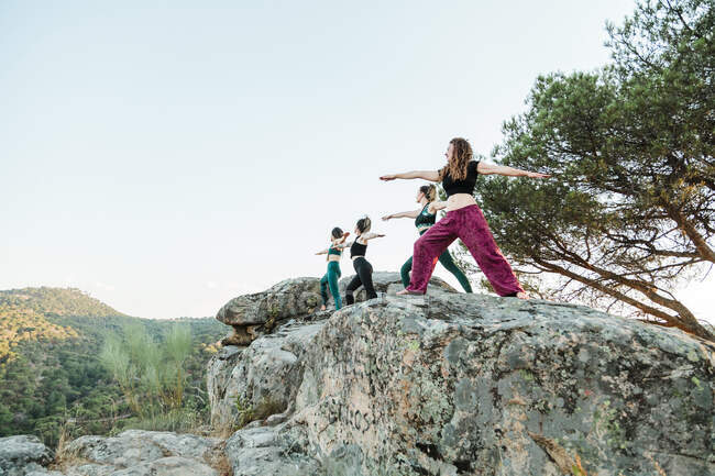 Mujeres amigas practicando yoga sobre rocas contra el cielo despejado durante el fin de semana - foto de stock