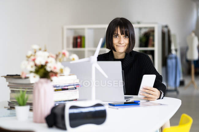 Молодая женщина с ноутбуком держит мобильный телефон во время учебы в кабинете — стоковое фото
