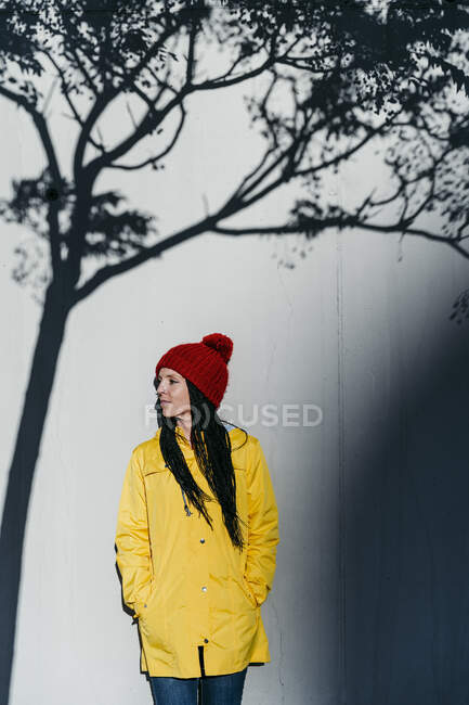 Woman wearing raincoat standing with hands in pockets under tree shadow against wall — Stock Photo