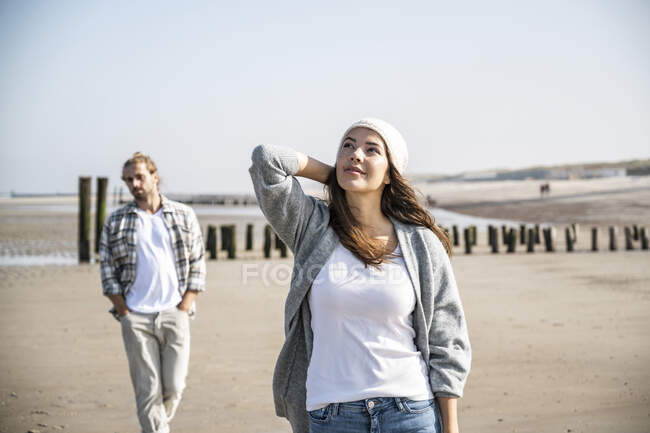 Thoughtful young woman looking up while man walking in background at beach — Stock Photo