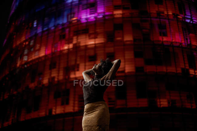 Женщина с поднятыми руками стоит против красного освещенного здания в городе — стоковое фото