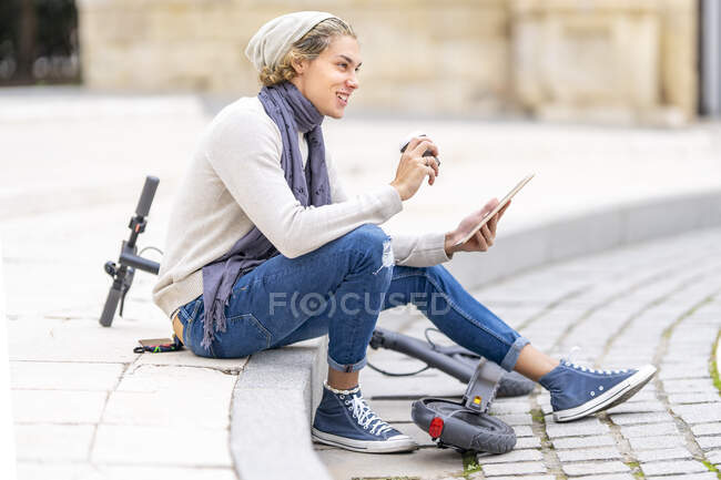 Uomo sorridente con tablet digitale bere caffè mentre seduto da scooter elettrico spinta in città — Foto stock