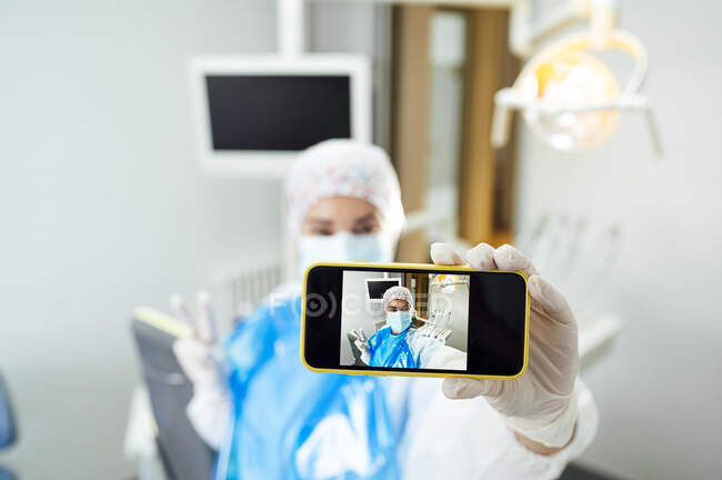 Стоматолог у захисному робочому одязі бере селфі через мобільний телефон, перебуваючи в клініці під час Ковід-19. — стокове фото
