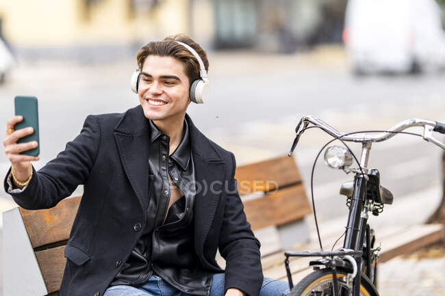 Sorridente bel giovane che si fa selfie attraverso lo smart phone mentre è seduto in bicicletta in città — Foto stock