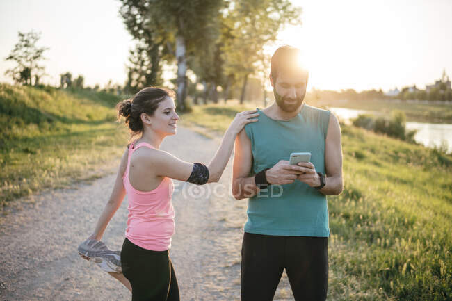 Männlicher Athlet benutzt Smartphone, während Sportlerin bei Sonnenuntergang mit erhobenem Bein im Park steht — Stockfoto