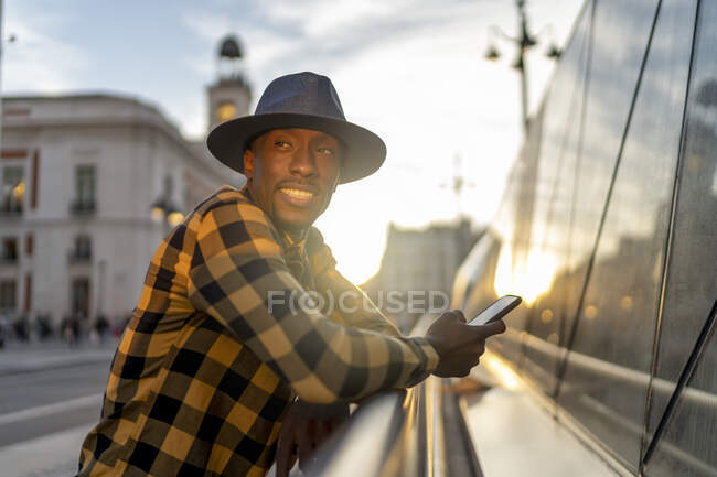 Hombre africano sonriente con teléfono inteligente mirando hacia otro lado mientras está de pie en la ciudad durante la puesta del sol - foto de stock