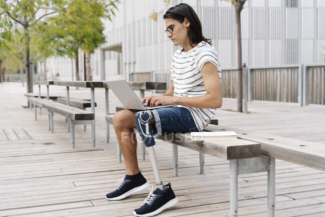 Contemplar al hombre con extremidad artificial usando el ordenador portátil mientras está sentado en el banco de la ciudad - foto de stock