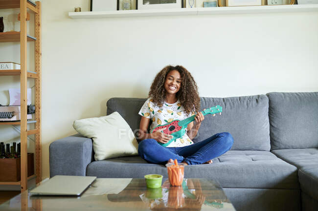 Smiling young woman with ukulele sitting on sofa in living room — Stock Photo