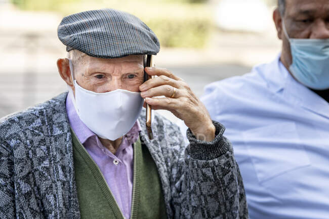 Homme âgé parlant sur un téléphone intelligent portant un masque protecteur assis avec un homme mature pendant la COVID-19 — Photo de stock