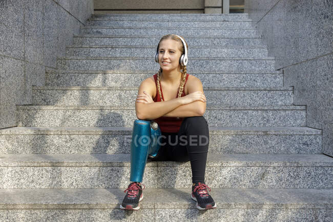 Femme sportive souriante avec une jambe prothétique portant des écouteurs assis avec les bras croisés sur l'escalier — Photo de stock