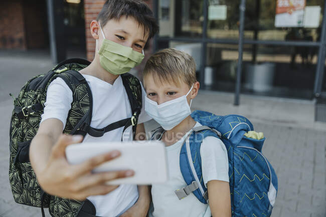 Frères prenant selfie sur téléphone intelligent tout en portant un masque protecteur debout devant le bâtiment de l'école — Photo de stock