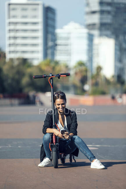Young woman using mobile phone while sitting on push scooter in city — Stock Photo