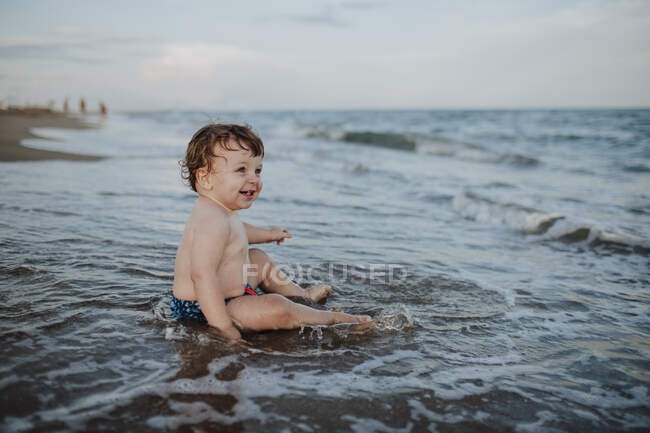 Niño disfrutando sentado en el agua en la playa al atardecer - foto de stock
