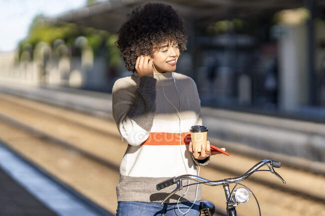 Mujer joven sonriente con bicicleta sosteniendo taza reutilizable mientras disfruta de la música en la estación de tren - foto de stock