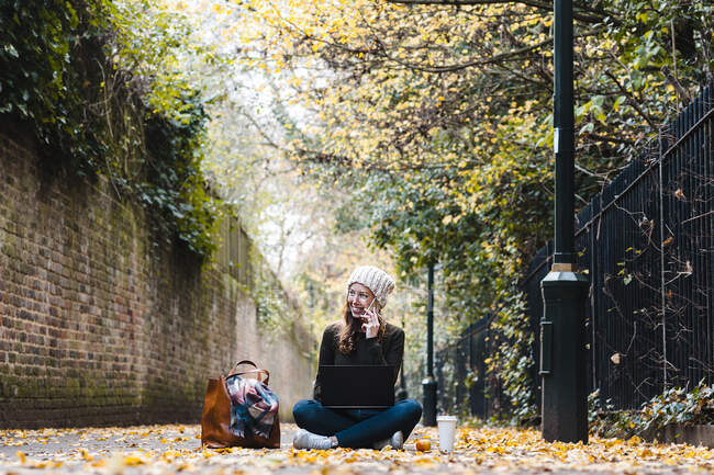 Smiling woman with laptop talking on smart phone while sitting on road — Stock Photo